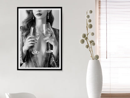 Framed artwork of a woman holding wine glasses on a wall next to a white vase with dried flowers.