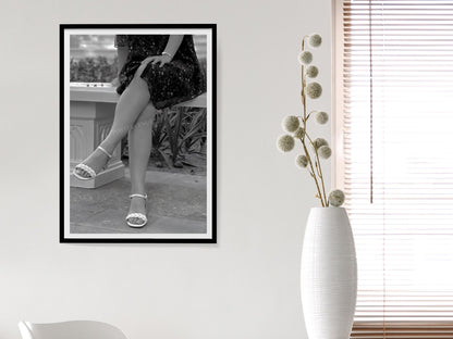 Framed black and white photo of a person in high heels on a wall next to a white vase with dried flowers.